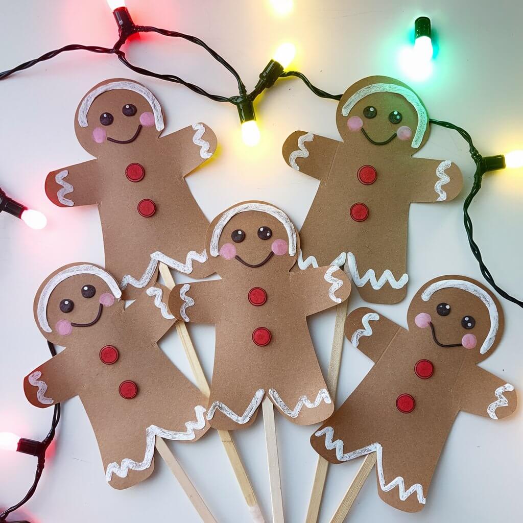 A holiday-themed play area with colorful lights and gingerbread men puppets. The puppets are created from brown cardstock and have expressive faces drawn on in detail. Each puppet is attached to a popsicle stick, allowing children to hold and manipulate them easily. The puppets are poised for storytelling, making this craft both decorative and interactive.