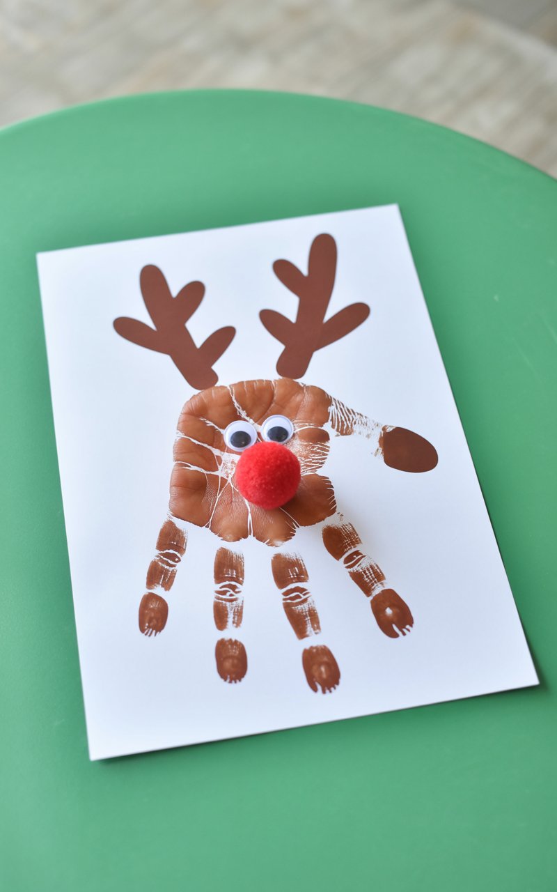 A photograph of a charming handprint reindeer craft displayed on crisp white paper against a plain green table surface. The reindeer is created from a brown painted handprint, with the thumb forming the head and fingers extending upward as antlers. Two white googly eyes are glued onto the thumb section, while a bright red pom-pom serves as the cheerful nose. The craft shows the delightful imperfections of a child's artwork, with slightly uneven paint coverage and endearing finger smudges around the edges.