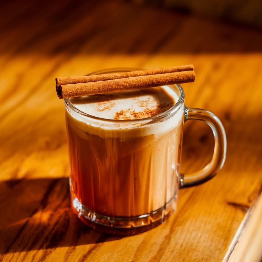 A photo of a hot buttered rum drink in a clear glass mug with a sturdy handle, placed on a warm-toned wooden table. The drink is a rich amber-brown color with a foamy layer on top. Two neatly stacked cinnamon sticks rest across the rim of the mug. The lighting is warm and natural.