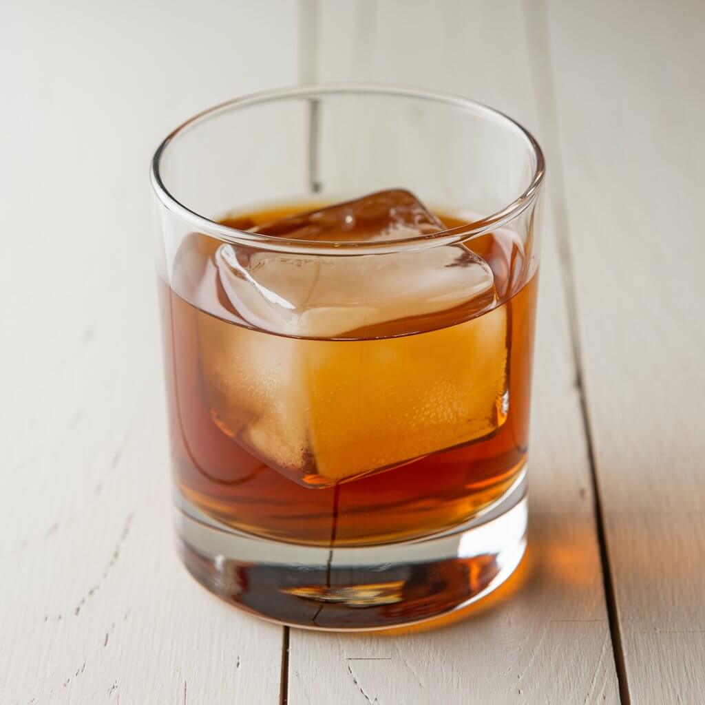 A photo of a classic maple old fashioned cocktail in a tumbler glass. The drink has a deep amber color and is served with a large ice cube. The cocktail is set on a bright white wooden table.