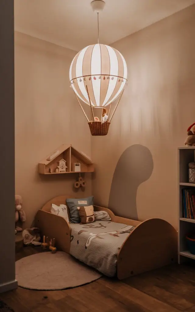 A photo of a small toddler bedroom with a fun pendant light shaped like a hot air balloon. The room has a cozy toddler bed design beneath the light. The walls are painted in a soft hue. There are a few toys and a bookshelf in the room. The flooring is wooden. The overall ambiance of the room is warm and inviting.