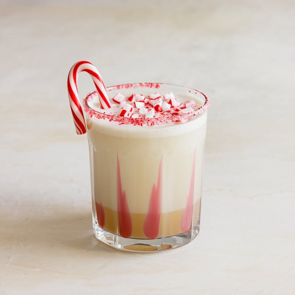 A peppermint white Russian cocktail in a glass with a creamy white base and a touch of pink peppermint swirl, topped with crushed candy canes. The glass has a candy cane rim and sits on a plain white table.