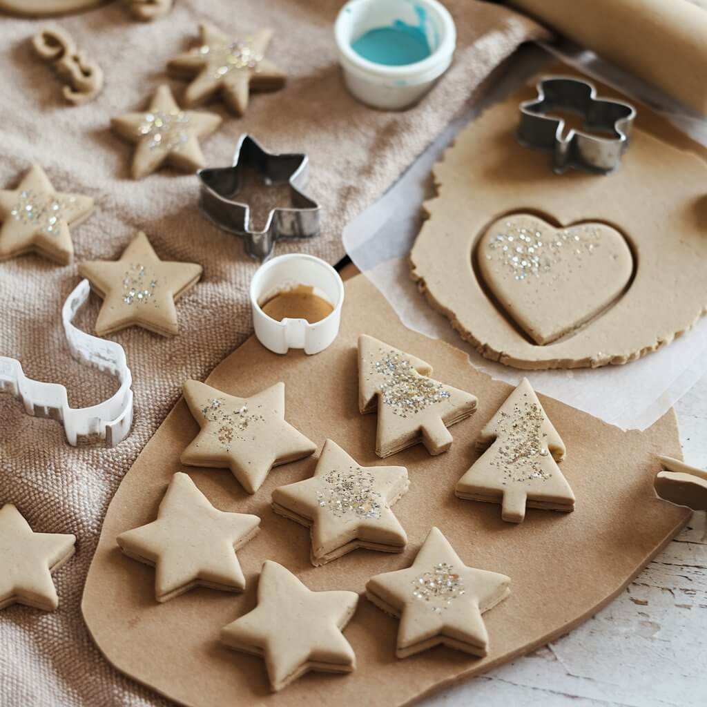 A photo of a craft setting with a collection of handcrafted salt dough ornaments. There are stars, trees, and hearts, all painted and decorated with glitter. There is dough being rolled out with cookie cutters. There are small bowls of paint. The background has a beige towel and a wooden surface.