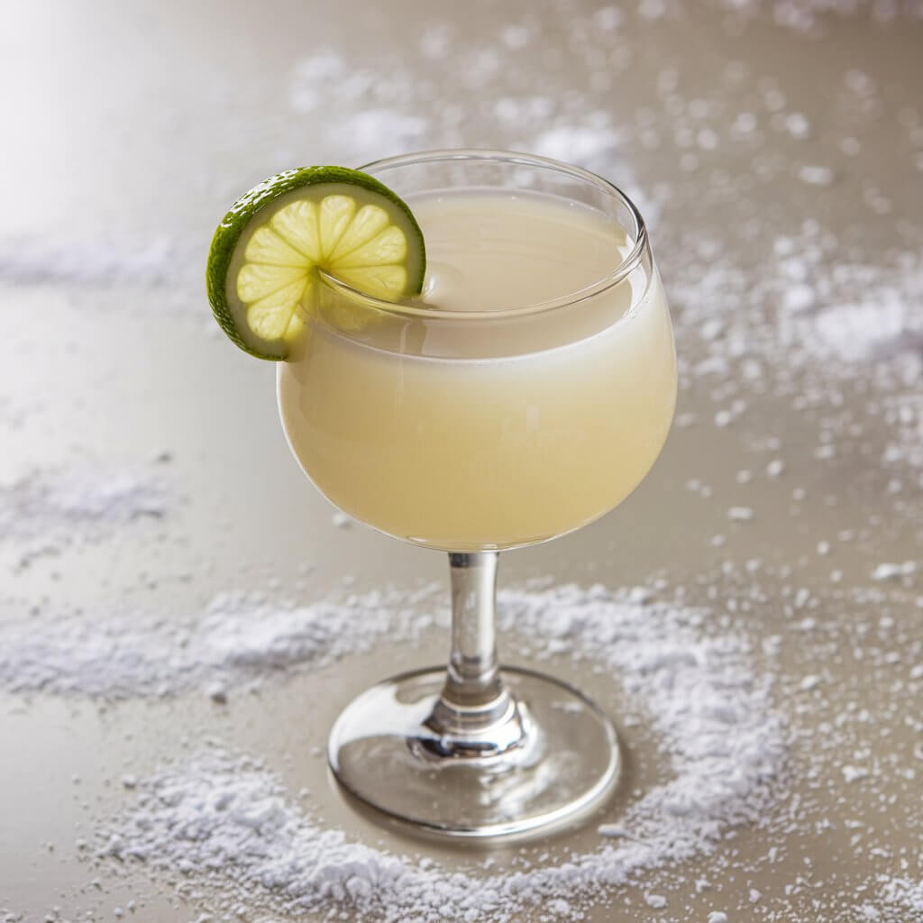 A retro snowball cocktail in a short glass, filled with a pale yellow creamy drink, topped with a lime wedge. The glass sits on a festive table scattered with snow-like powder. The background is a bright white table.