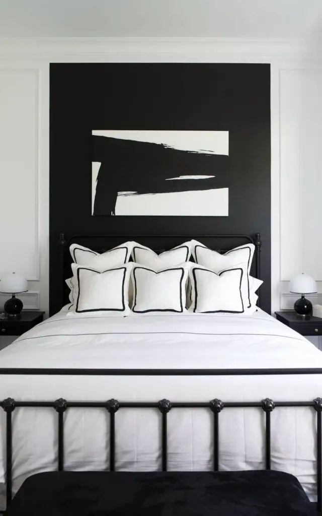 A black and white bedroom with a bold black accent wall behind a cozy bed draped in luxurious white linens. The black wall contrasts sharply with the white walls around it, giving depth to the space. The bed features black metal frame detailing and white pillows with black trim, creating harmony in the color scheme. A large, modern black and white painting hangs above the bed, adding visual interest, while a small black nightstand with a white lamp completes the stylish look.