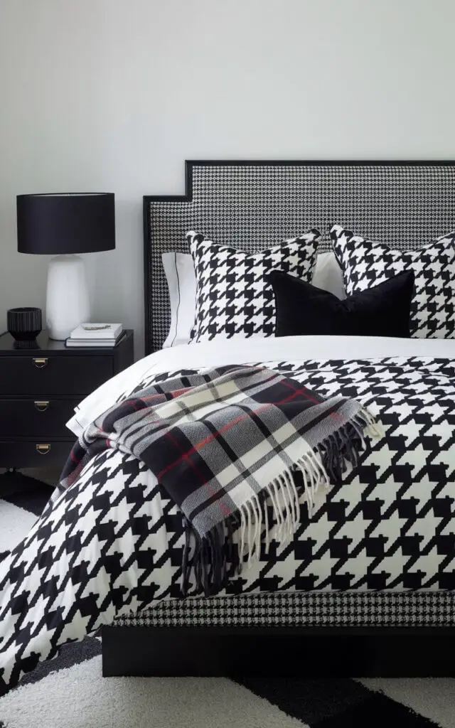 A black and white bedroom with a high-contrast houndstooth pattern on a very cozy bed. The bedding features bold black and white houndstooth, while a plaid throw in similar colors lies at the foot. A black nightstand with a white lamp adds balance, creating a dynamic and layered aesthetic.