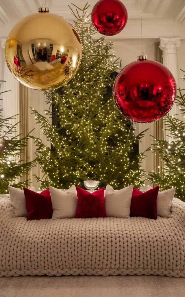 A spacious Christmas living room beautifully decorated with oversized ornaments hanging from the ceiling and placed strategically around the room. These large, shimmering ornaments in gold and red create a stunning visual impact against the backdrop of a very cozy sofa draped with a thick, knitted blanket. The room is further enhanced with a grand Christmas tree and sparkling lights, making it a perfect festive retreat.