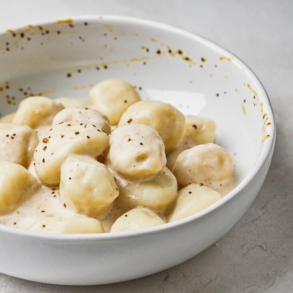 A close-up view of a white, shallow ceramic bowl filled with gnocchi coated in a creamy, white sauce. The gnocchi pieces are plump, slightly irregular in shape, and lightly coated with a sauce that has a smooth, glossy texture. Black pepper specks are scattered throughout the sauce, adding a subtle contrast to the creamy color. Some of the sauce has splattered along the inside rim of the bowl, as well as outside, giving a casual, imperfect look to the presentation. The bowl is placed on a light gray surface. The lighting is natural and soft, emphasizing the richness of the sauce and the pillowy texture of the gnocchi. The overall composition is minimalist, focusing on the gnocchi and sauce without any additional garnish or background elements.