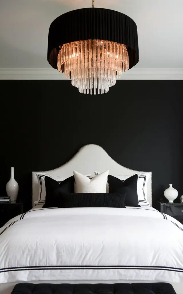 A dramatic black and white bedroom with a bold black chandelier above a very cozy bed with white linens. The chandelier adds sophistication and elegance, drawing attention upward. The bed is accented with black and white pillows, and a small black nightstand with a white vase completes the luxurious look of this stunning bedroom.