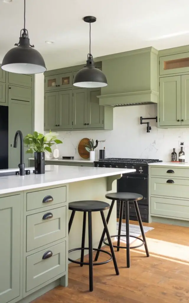 A photo of a modern sage green kitchen with striking black accents. The sage green cabinets are fitted with matte black handles, and black bar stools sit at the kitchen island. Overhead, black pendant lights provide illumination, while the black hardware contrasts against white marble countertops, creating a dynamic visual impact.