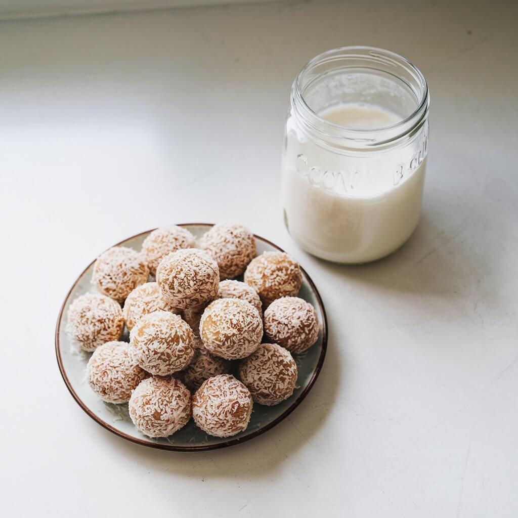 A photo of a small plate of round coconut milk energy bites, coated in tiny shredded coconut and displayed on a bright white kitchen countertop. The energy bites have a cream color and are arranged in a circular pattern. A half-empty jar of coconut milk sits nearby, showcasing the wholesome ingredients used in making the energy bites. The background is clean and minimalistic.