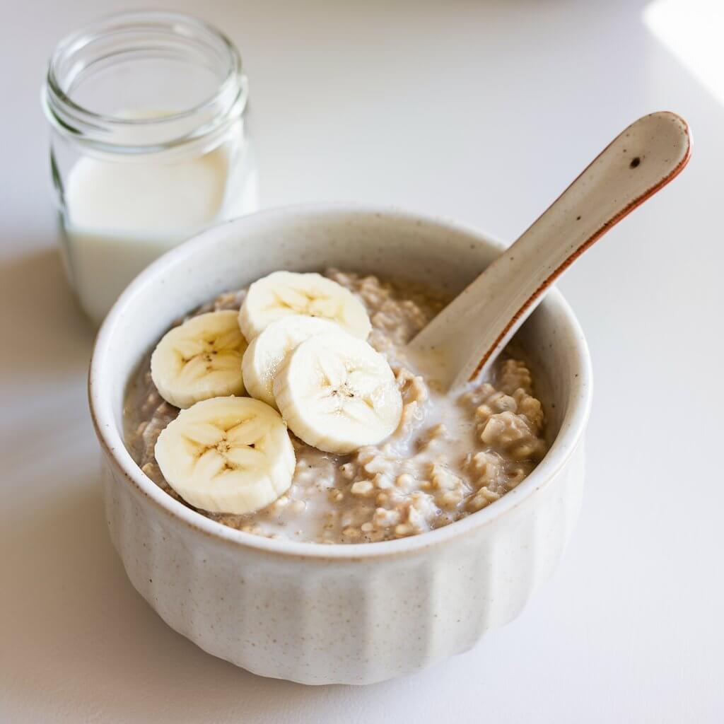 A photo of a hearty bowl of white ceramic bowl of coconut milk oatmeal topped with sliced bananas. A ceramic spoon is dipped into the bowl, and a small glass jar of coconut milk is placed beside the bowl. The setting is a bright plain white countertop.