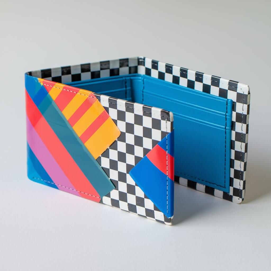 A photo of a homemade duct tape wallet crafted by a kid. The wallet features vibrant, checkered stripes duct tapes with smooth, homemade looking clean edges. The wallet is showing an organized arrangement of card slots and a bill compartment. The wallet is covered with duct tape. The design includes a glossy finish that reflects light subtly, emphasizing the tape's quality. The wallet is placed on a plain white table, offering a sharp contrast to highlight the vibrant colors and the precise craftsmanship of the wallet.