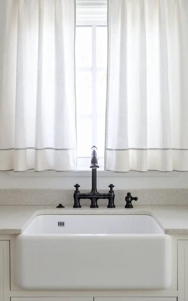 A minimalist farmhouse kitchen sink with a sleek black faucet. Above the sink, there are plain white cotton kitchen window curtains. The curtains are hemmed with subtle gray stitching for a touch of elegance. The overall simplicity pairs well with polished granite countertops, creating a clean and modern farmhouse look.