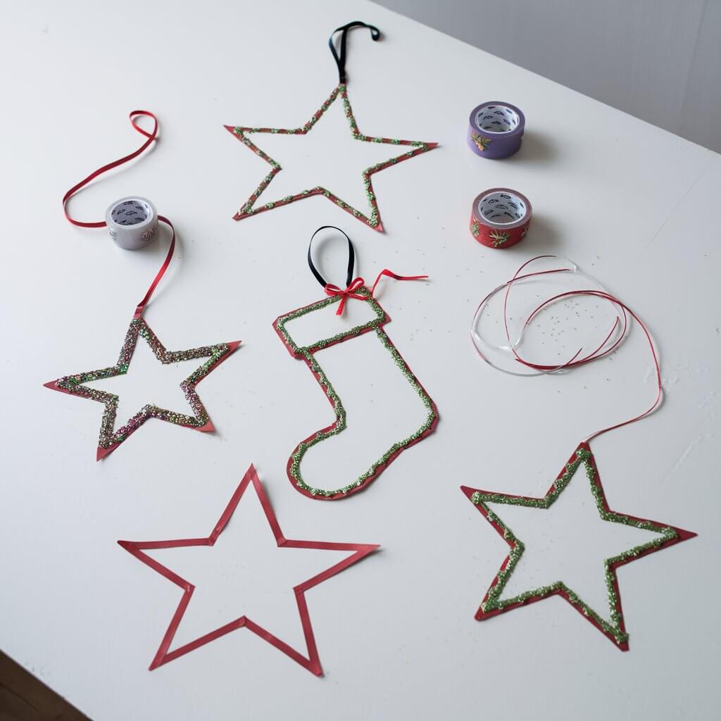 A collection of homemade duct tape Christmas ornaments on a plain white table. There are thin stars, a stocking, and a thin light bulb made from plain duct tape. Each ornament is detailed with tiny embellishments like glitter or bows. They have delicate loops of ribbon. There are duct tapes on the table. The vivid colors and festive designs stand out sharply against the clean background.