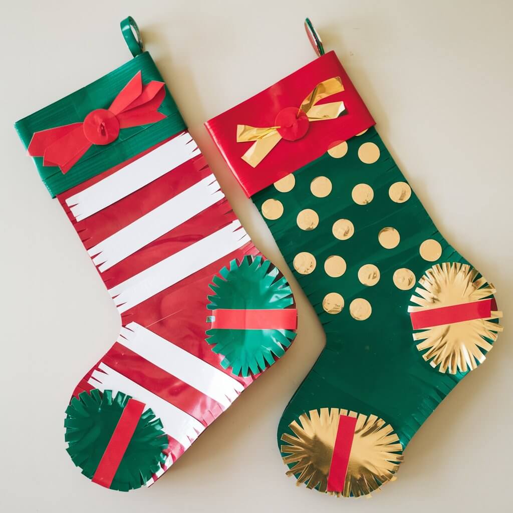 A photo of a pair of homemade duct tape Christmas stockings crafted by kids. One stocking is red with white stripes and the other is green with gold accents. Both stockings have neatly sealed seams and are large enough to hold gifts. The duct tape is visible on the craft. The stockings are laid flat on a plain white table. The vibrant colors and cheerful designs add holiday charm to the scene.