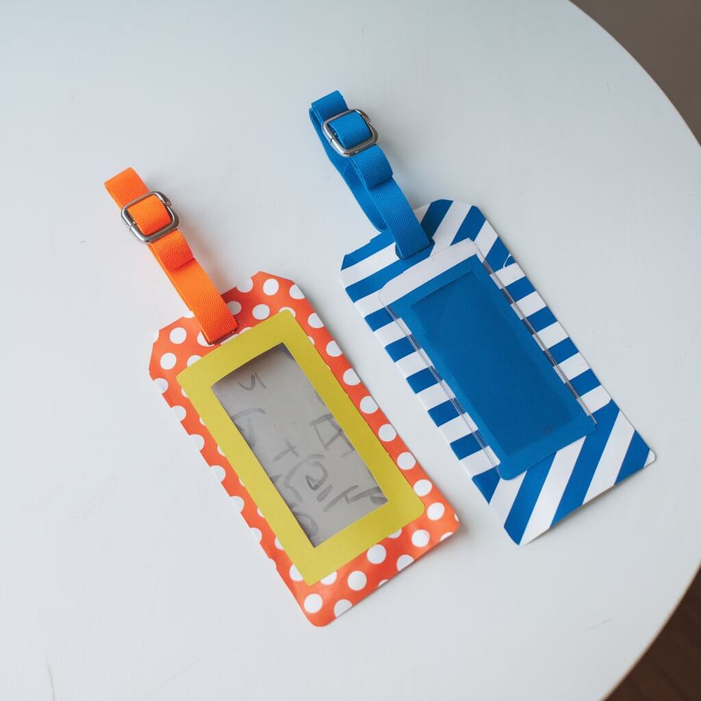 A photo of two homemade duct tape luggage tags on a white table. One tag is bright orange with polka dots, and the other is blue with stripes. Each tag has a clear window for contact information and a sturdy strap for attaching to luggage. The background is plain white.