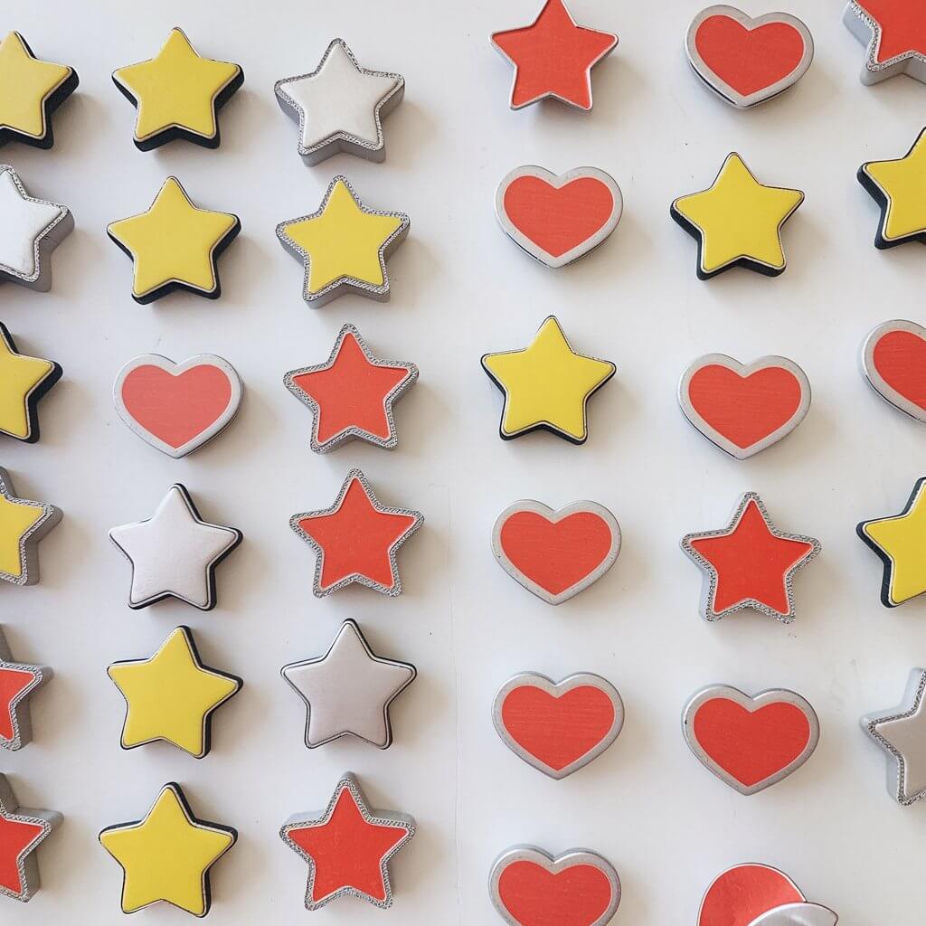 A photo of a plain white table with a variety of homemade duct tape magnets. There are stars, hearts, and circles in bright colors like yellow, red, and silver. Each magnet has a smooth finish and a strong magnetic backing for functionality. The stars, hearts, and circles are arranged next to each other. The background is clean and simple, allowing the shapes and vibrant colors of the magnets to pop.