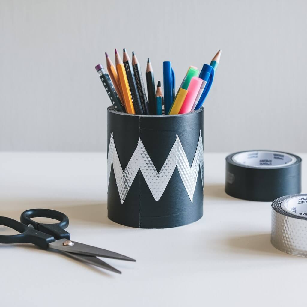 A photo of a cylindrical pencil holder made from black duct tape, decorated with a shiny silver zigzag design. The holder is filled with colorful pencils and pens, placed on a plain white table. A pair of scissors, a roll of black duct tape, and a roll of silver duct tape sit nearby, showing the tools used.