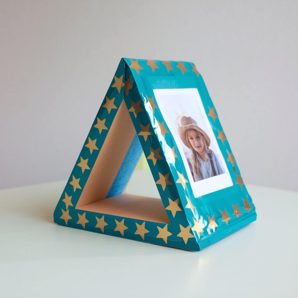 A photo of a kid's homemade craft of a duct tape picture holder with a triangle-shaped base. The holder is covered with bright teal duct tape and decorated with gold stars. The holder securely displays a small photo and is compact enough for a desk or shelf. The craft is placed on a plain white table, and the glossy texture and vibrant colors of the duct tape shine prominently, enhancing the creative design.