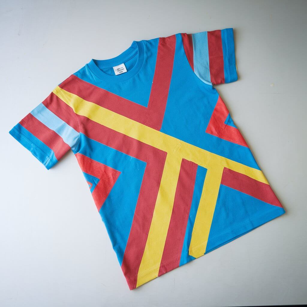 A homemade duct tape t-shirt with a bold graphic design of colorful stripes and shapes in shades of red, blue, and yellow. The duct tape is carefully adhered to the fabric for a smooth, wrinkle-free finish. The t-shirt is laid flat on a plain white table. The vibrant colors and striking geometric patterns draw attention, highlighting the creativity behind this wearable art. The craft was made by a kid.