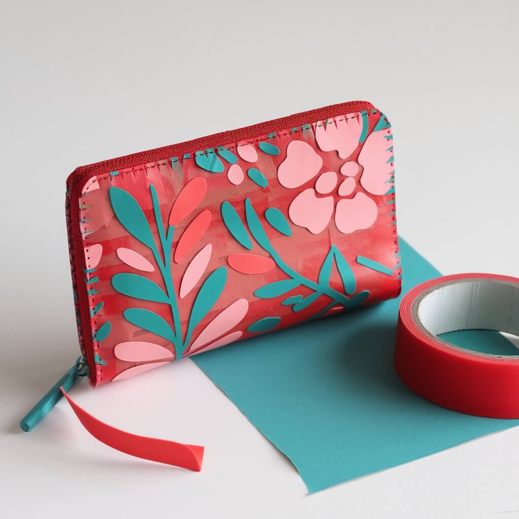 A photo of a homemade small duct tape wallet purse covered with duct tape. The purse has a vibrant floral duct tape design in red and pink and a teal duct tape is also used. It is complete with a functional zipper and strap. The edges are carefully sealed for durability, and the overall shape is sleek and modern. The purse shows the cuts of duct tape lines. Positioned on a plain white table, the bright design contrasts beautifully against the backdrop, showcasing every intricate detail of this stylish accessory. A big red duct tape is on the table matching the purse and teal duct tape.