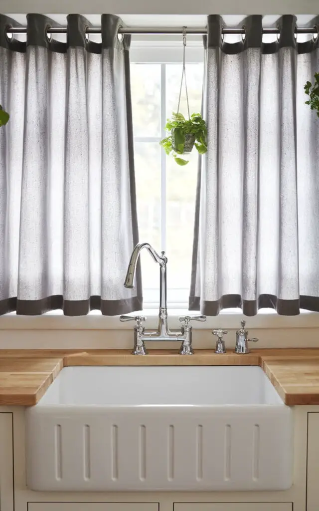 A photo of a farmhouse kitchen sink with a polished stainless-steel modern faucet. The sink is placed under a simple gray grommet-style kitchen window curtains. The curtains have evenly spaced pleats, giving a clean, modern look. Above the kitchen sink, there are natural wood countertops and hanging herb pots, adding warmth and functionality to the space.