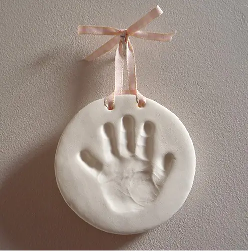 A smooth, round clay keepsake featuring the pressed handprint of a child at its center. The off-white clay has a polished finish, highlighting the fine details of the handprint, including the delicate lines of the fingers and palm. Two small holes near the top are threaded with a soft pink ribbon tied into a neat bow, allowing the keepsake to be hung. The design is simple and elegant, showcasing the timeless beauty of this personalized memento against a neutral background.