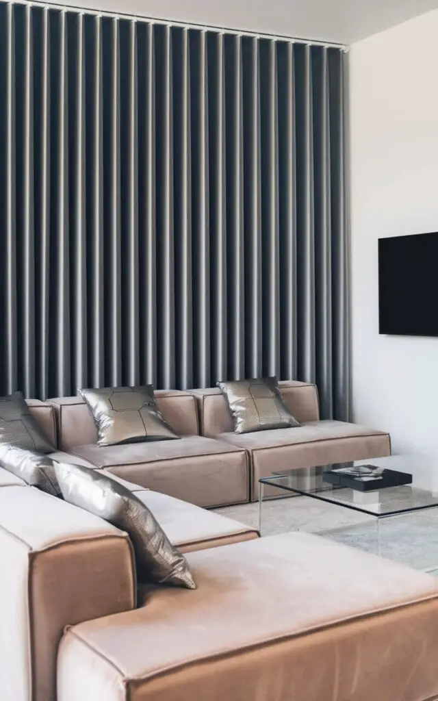 A photo of a futuristic living room with a charcoal gray motorized curtain that opens with a remote control. There's a cozy modular sofa in soft beige with tech-inspired metallic pillows. A glass coffee table and a smart TV mounted on the wall complete the high-tech aesthetic.