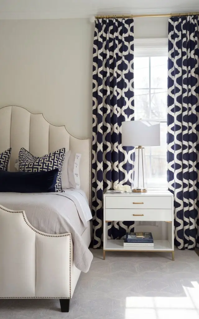 A photo of a cheerful bedroom with a bold, geometric patterned navy and white window curtains. The curtains create a striking focal point, contrasting beautifully with the light-colored walls. A very cozy upholstered bed with a scalloped headboard in a plush velvet fabric stands nearby, dressed in neutral bedding with coordinating navy throw pillows. A white bedside table with gold hardware sits beside the bed, paired with a stack of books and a sleek, modern lamp.