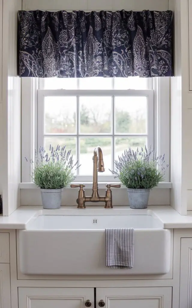 A photo of a white farmhouse kitchen sink with a bronze pull-down faucet. The sink is framed by a patterned valance-style kitchen window curtain above it. The curtain showcases a navy blue paisley design, adding a touch of elegance. White paneled cabinetry and potted lavender plants complete the sophisticated look.