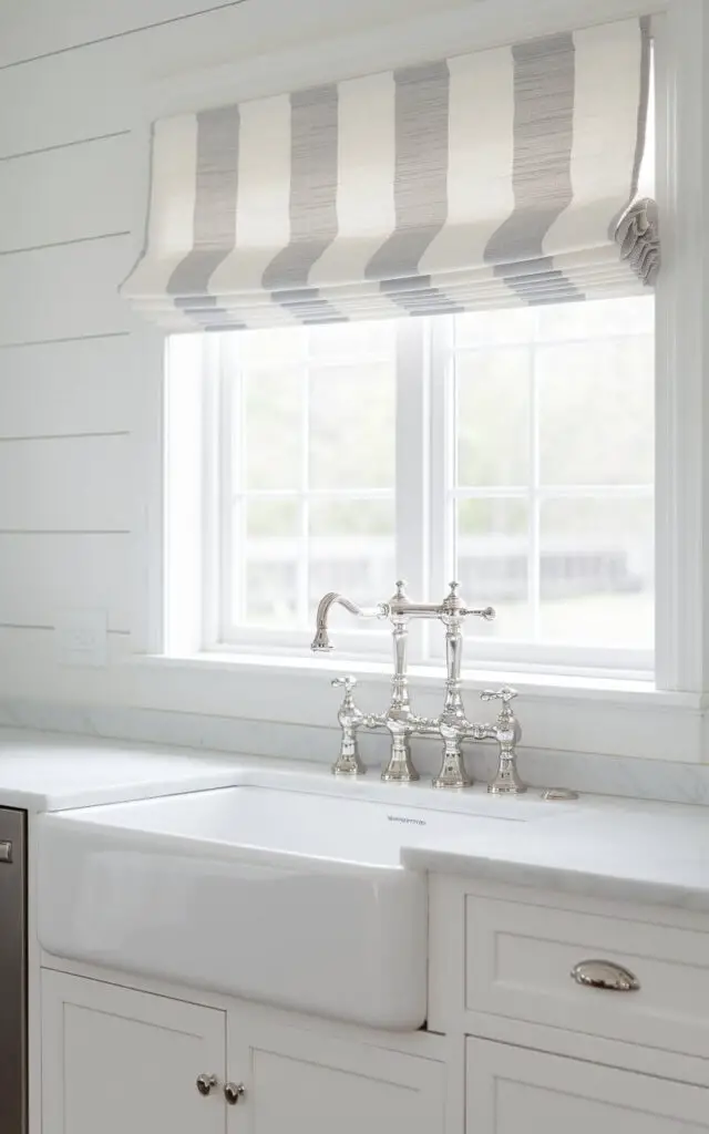 A bright farmhouse kitchen with a polished stainless-steel faucet and a crisp white farmhouse kitchen sink. Above the kitchen sink, there is a Roman shade-style kitchen window curtain. The curtain has a subtle striped pattern in soft gray and white, perfectly matching the shiplap walls and marble countertops.