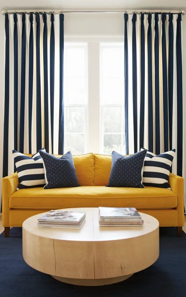 A bright and dynamic living room with curtains featuring bold navy and white vertical stripes. A very cozy yellow sofa adds a cheerful touch, layered with striped and polka-dot pillows. A round coffee table with a natural wood finish hosts a stack of magazines, and the navy area rug ties the look together.