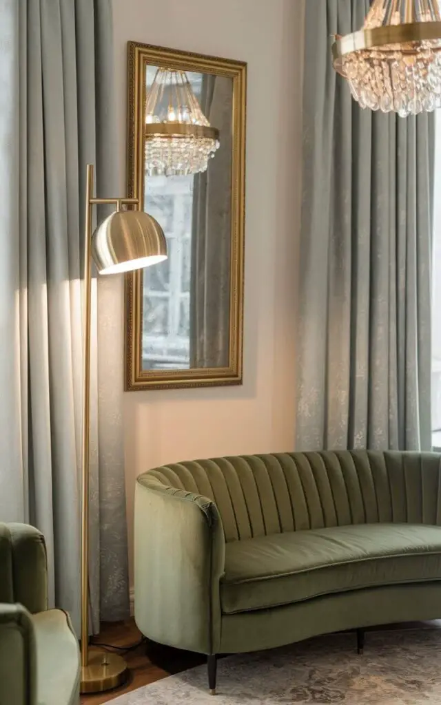 A photo of an elegant living room with a green couch and brass lighting. There is a tall brass floor lamp beside the couch and a brass chandelier hanging overhead. The light reflects off a gold-framed mirror on the wall, creating a luxurious ambiance. Soft gray curtains and a patterned rug are also present, completing the refined look.