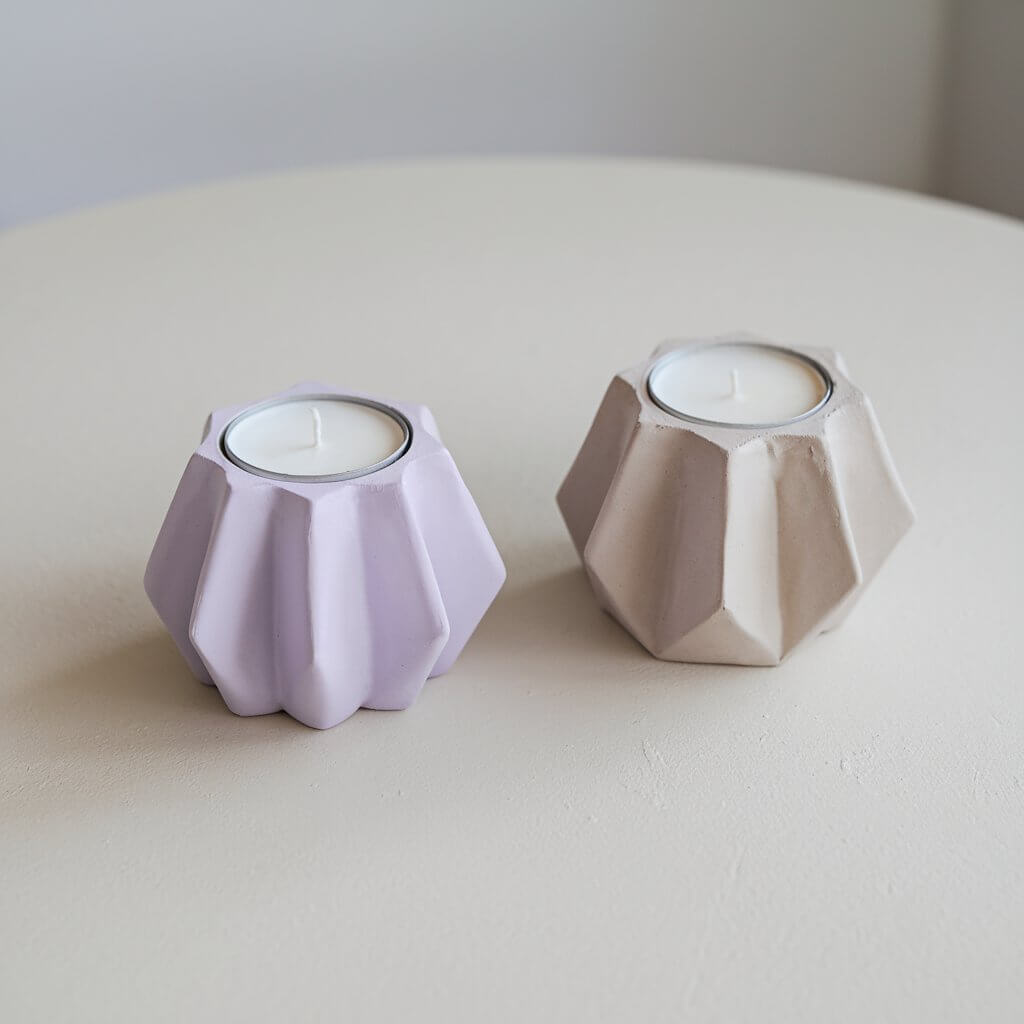 A photo of two homemade, simple-looking clay candle holders on a plain white table. The candle holders are shaped like elegant geometric designs. One holder is painted in soft lavender, while the other has a natural gray clay finish. Both holders have grooves to securely hold candles and a smooth surface for a polished appearance.