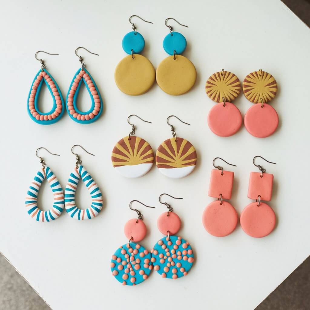 A chic collection of homemade simple-looking clay earrings in trendy shapes like teardrops, circles, and geometric designs. They are placed on a plain white table. Each pair is painted in bold colors like turquoise, mustard yellow, and coral, with metallic hooks attached. Some earrings feature intricate patterns, while others have simple, solid colors.