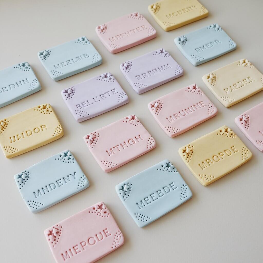 A photo of a collection of homemade, simple-looking clay name tags placed neatly on a plain white table. Each tag is rectangular and features a name etched into the surface using small stamps. There are designs like tiny flowers and stars embossed on the corners. The clay tags are painted in soft pastel shades of blue, pink, and yellow. Some of the tags are left unpainted for a natural look.