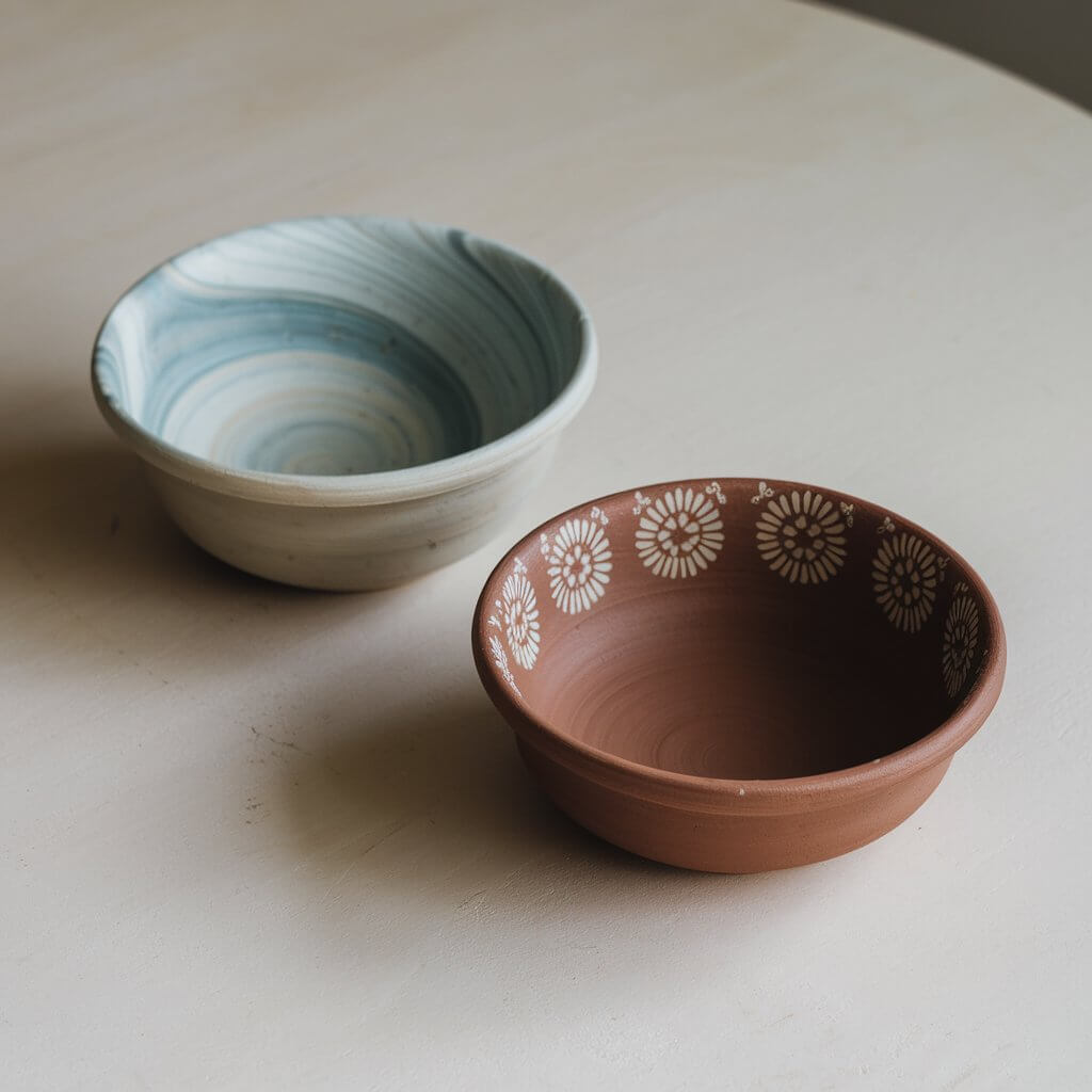 A photo of two medium-sized homemade, simple-looking clay bowls. One bowl has a marble-like swirl of blue and white, while the other is a solid brown color with intricate floral patterns painted on its rim. The bowls have smooth, rounded edges and a slightly matte finish, indicating their handmade origin. They are placed on a plain white table.