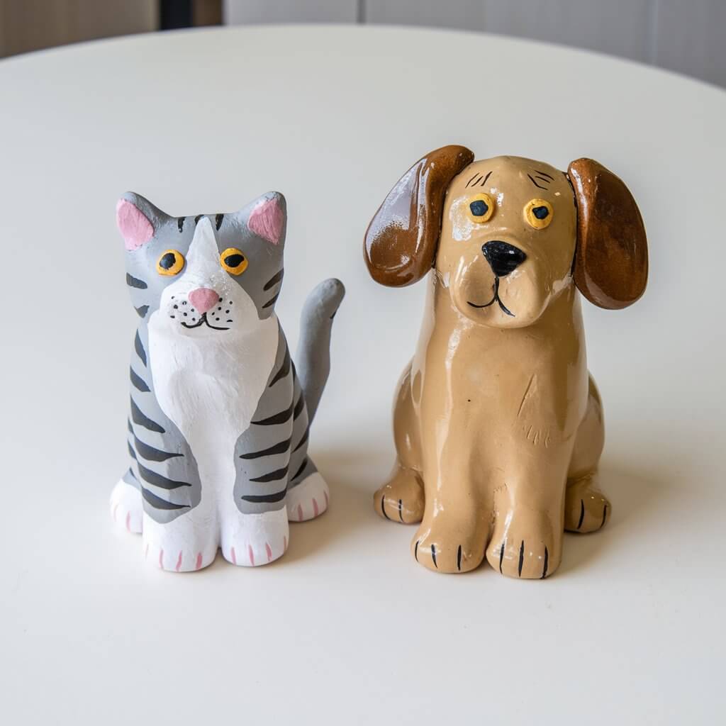 A photo of a plain white table with two homemade, simple-looking clay pet figurines. There's a cat with painted gray and white stripes and a dog with floppy ears and a glossy golden coat. Both figurines have detailed features like paws, whiskers, and tails. The background is clean and uncluttered.A photo of a plain white table with two homemade, simple-looking clay pet figurines. There's a cat with painted gray and white stripes and a dog with floppy ears and a glossy golden coat. Both figurines have detailed features like paws, whiskers, and tails. The background is clean and uncluttered.