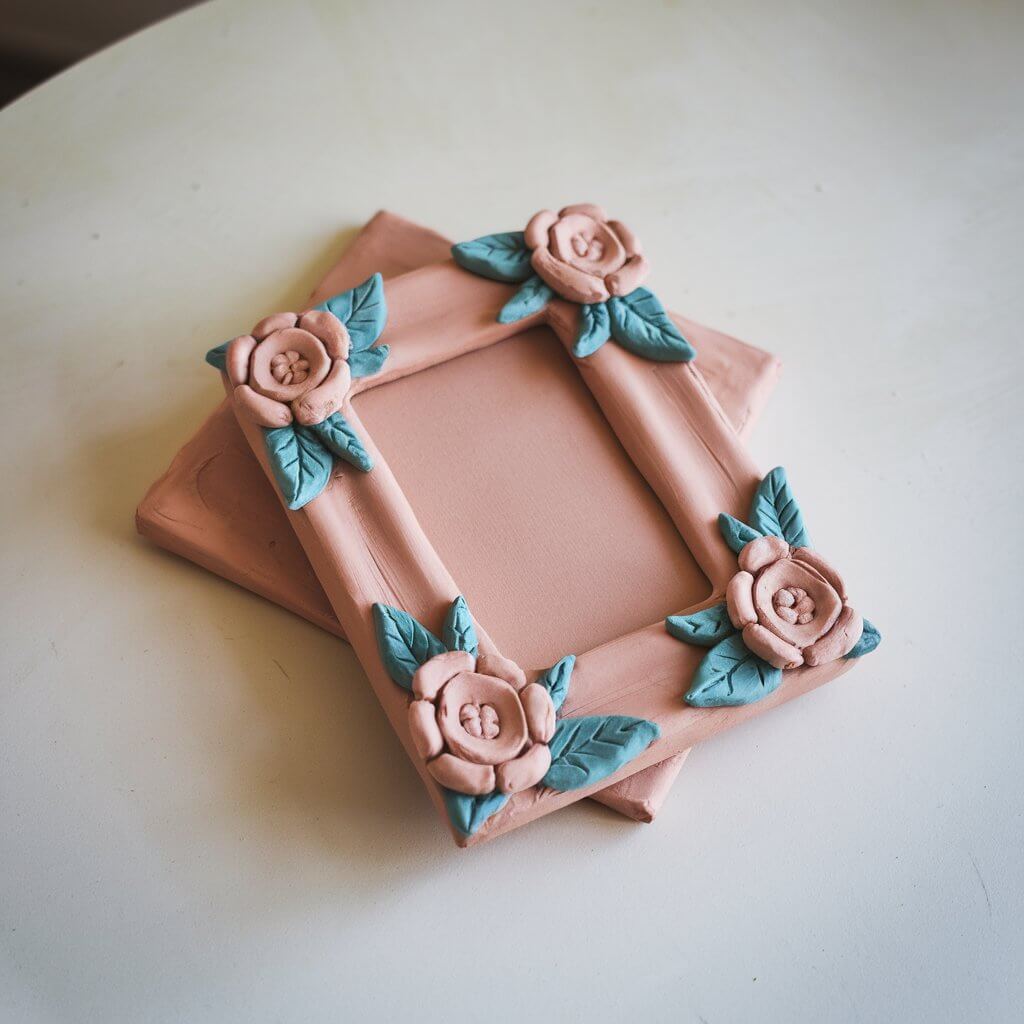 A photo of a homemade clay picture frame. The frame has a rectangular shape and is resting on a plain white table. The frame features raised floral decorations on its edges, painted in soft pastels. The center is open for inserting a photo. The frame is made of clay, and its backing is also crafted from a separate piece of clay.