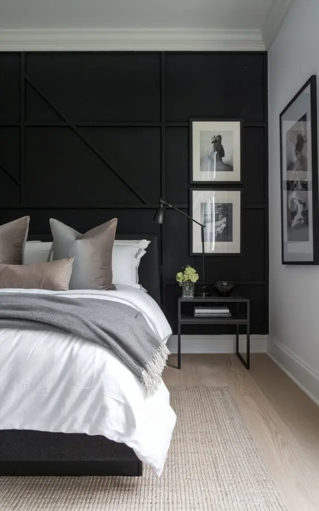 A photo of a dark bedroom with a dramatic black accent wall, contrasting beautifully with neutral-toned furnishings. The cozy bed is adorned with crisp white linens, gray throw blankets, and soft taupe pillows. The black wall is highlighted with framed monochrome art, while a modern bedside table holds a sleek reading lamp. A woven area rug adds texture to the light wooden floor, balancing the boldness of the dark wall.