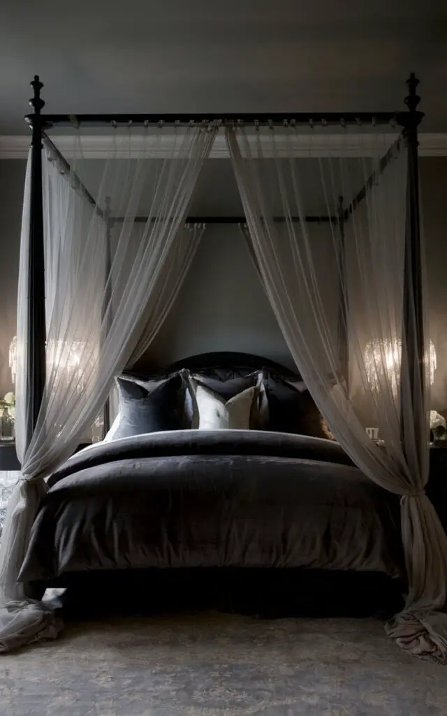 A photo of a luxurious canopy bed in a dark bedroom. The bed has flowing sheer drapes and dark gray bedding. The bed is framed by black wooden posts. There are bedside sconces that provide soft lighting. The walls are painted a soft charcoal color. There is a plush area rug beneath the bed.