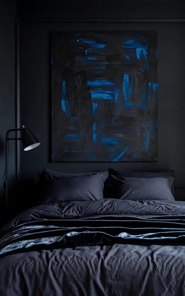 A dark bedroom with a bold, moody abstract painting in deep blues and blacks hanging above a cozy bed dressed in charcoal gray linens and a velvet throw. The surrounding walls are painted a matching dark tone, making the artwork stand out. A black-framed floor lamp adds both light and artistic appeal.