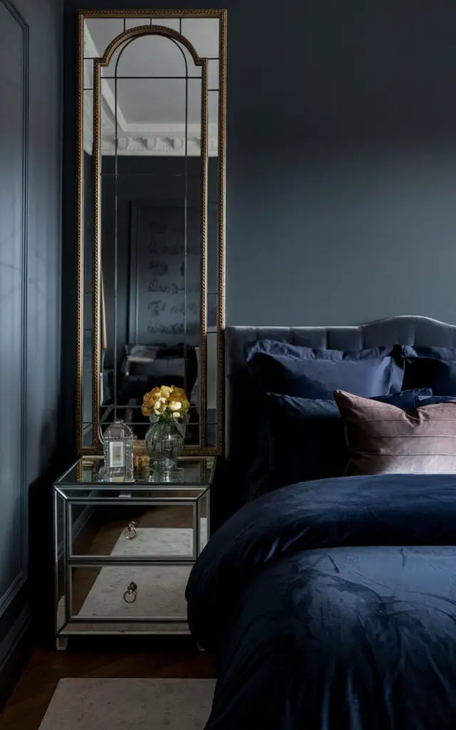 A photo of a dark bedroom with mirrors that amplify the light. There's a large, gold-framed mirror opposite a cozy bed with navy bedding and velvet pillows. The walls are painted a deep slate gray. A mirrored nightstand adds an extra touch of shine, enhancing the room's sophistication.