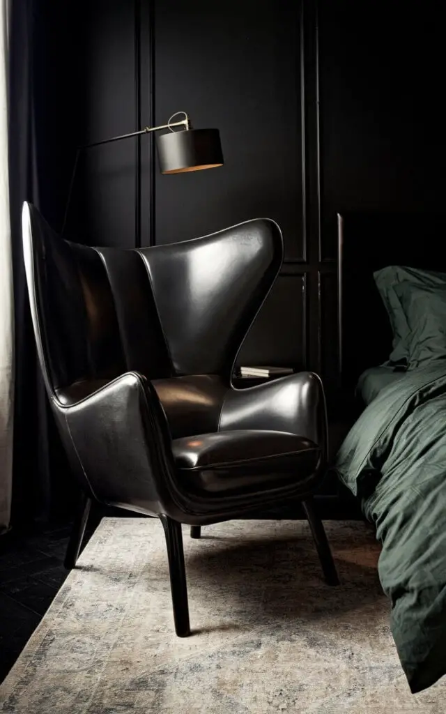 A dark bedroom with a bold statement chair near a cozy bed with deep green bedding. The chair has dark leather upholstery with sleek, modern lines. The black walls and an oversized floor lamp create a moody ambiance. A soft area rug ties the space together, and the chair provides a cozy nook for reading or relaxing.