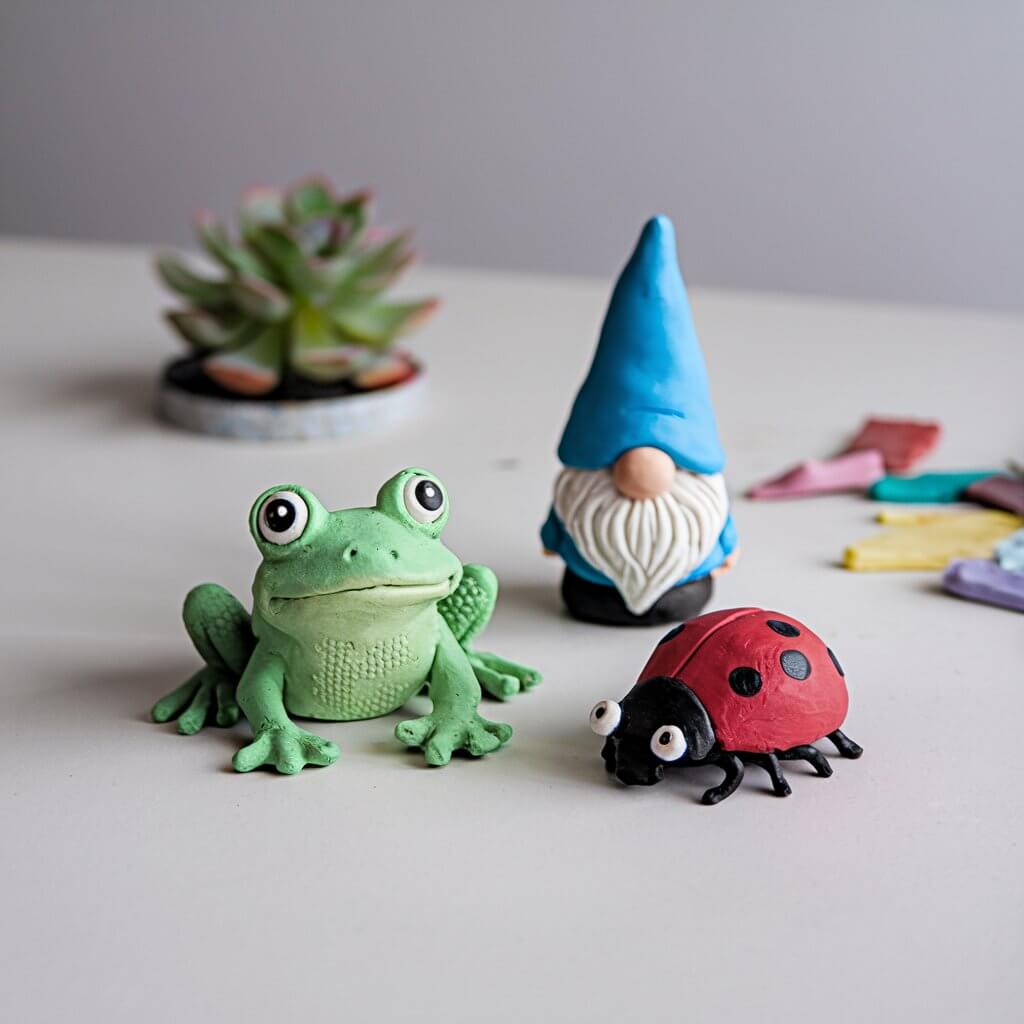 A photo of a set of miniature clay garden creatures. There's a charming green frog with large eyes and textured skin. There's a detailed red ladybug with multiple spots. There's a small gnome wearing a pointed blue hat. The creature are placed on a plain white table. In the background, there's a small succulent plant and scattered pieces of colorful clay.