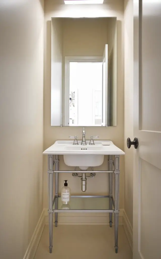 A photo of a small powder room with a vintage-inspired console sink. The sink has slender legs and clean lines, giving it an airy feel. The room has a modern design and is painted in soft ivory. The walls have a large square mirror above the sink, and the door is open, revealing an entryway. The room has a minimalist faucet with a sleek modern finish. A clear soap bottle is placed on the console's bottom shelf. The metallic accents of the sink and faucet stand out against the soft background.