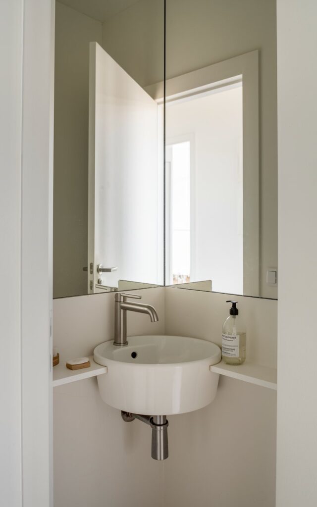 A photo of a small powder room with a corner sink. The room has a minimalist design with a white basin sink and a compact modern faucet in matte chrome. There is a clear soap bottle on a corner shelf. The room has a triangular mirror above the sink, which reflects the bright open white door in the background. The door leads to an entryway, visually extending the small footprint of the room.