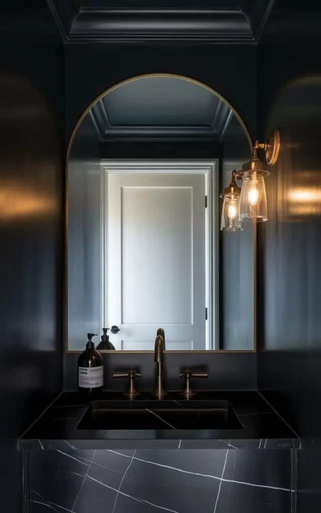 A photo of a small powder room with a deep charcoal palette. The walls, ceiling, and trim are painted the same sultry shade. A modern faucet in brushed bronze stands out against a black marble vanity. A sleek, black-labeled soap bottle and a large arched mirror, edged in brass, are also present. The room has a white door in the background. An antique sconce adds warmth and an ambient golden hue.