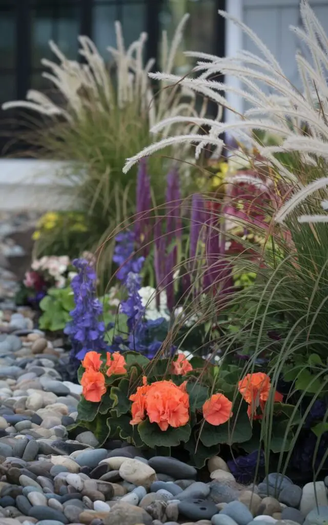 A photo of a flower bed in front of a house. The bed is decorated with various flowers, including begonias and salvias, and decorative grasses. The grasses add height and texture, and their plumes catch the light, creating motion in the space. The bed is bordered with river stones, adding a natural, flowing feel. The overall design has a modern yet relaxed feel.